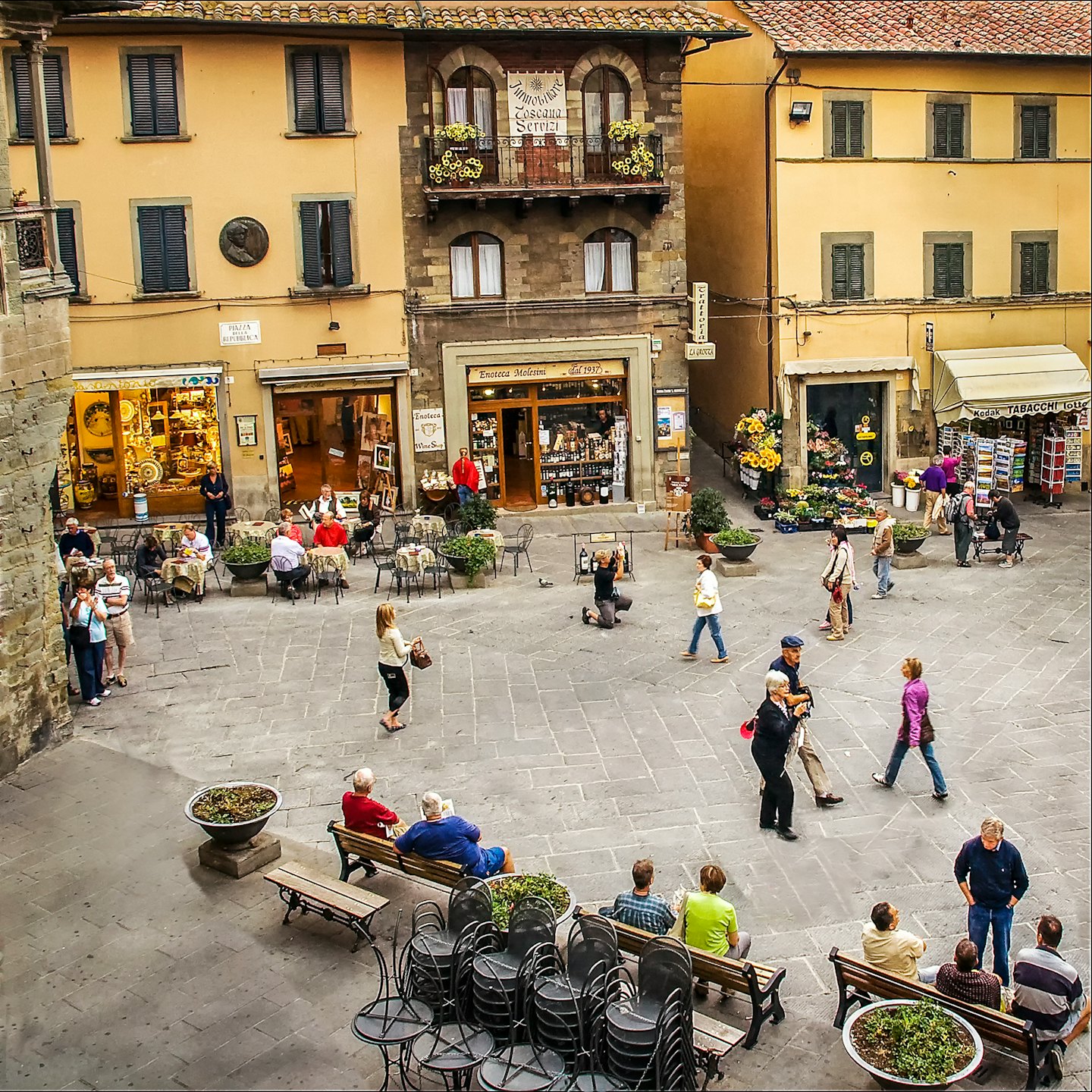 Persone passeggiano e siedono nei caffè in una piazza cittadina europea affollata.